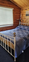 Una habitación de cabaña de madera con una cama, una ventana con persianas y una lámpara fijada a la pared.
