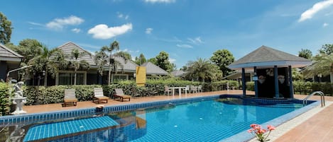 Una piscina con sillas de descanso y un pabellón, rodeada de vegetación y edificios.