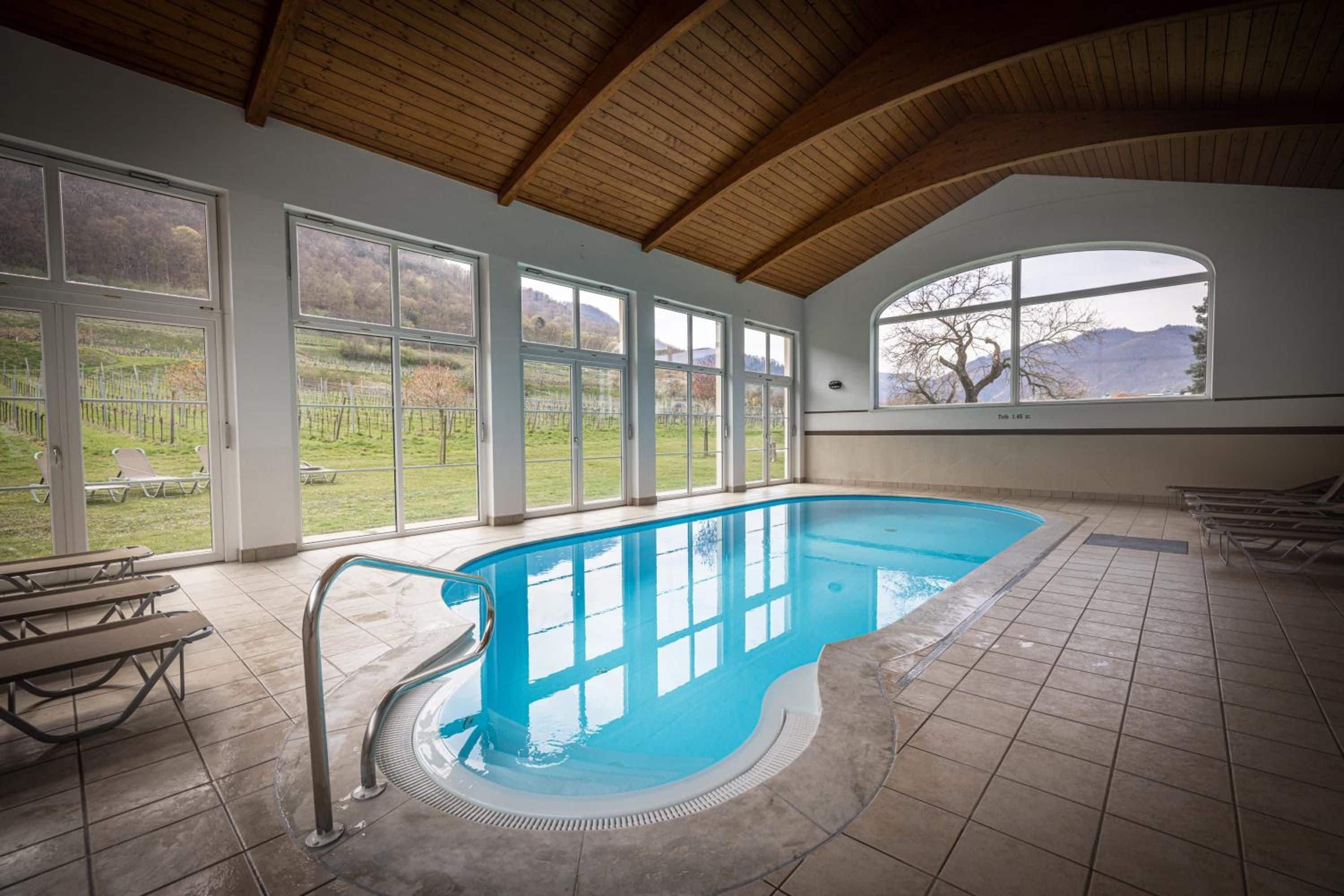 An indoor swimming pool with a wooden ceiling, large windows, and a view of a vineyard.