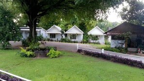 A row of white cottages with a thatched-roof building on the right, surrounded by greenery and a paved path. - Hadsaisuay Resort And Homestay (Kanchanaburi)