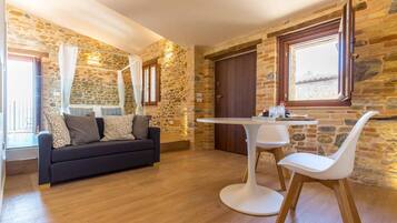 Ein Zimmer mit Steinwand, Holzboden, einer schwarzen Couch mit Kissen, einem weißen Tisch mit Stühlen und einem Fenster mit Vorhängen.