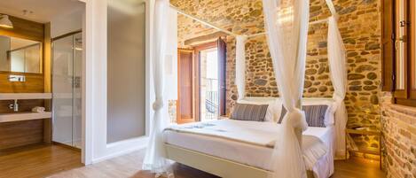 A bedroom with a stone wall, a four-poster bed, and a bathroom with a glass shower enclosure.