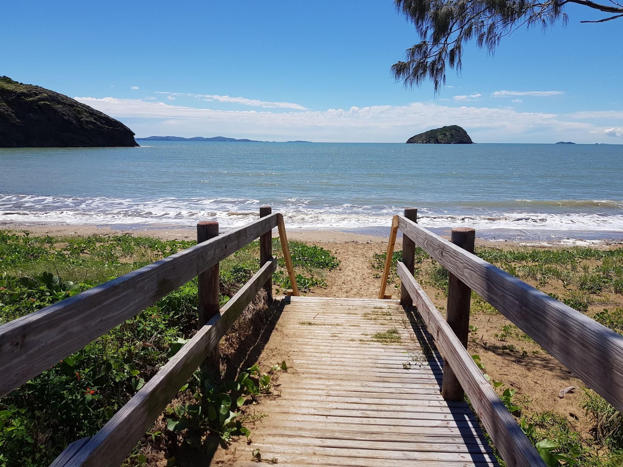 A wooden boardwalk leading to a beach with waves, a distant island, and a clear sky.