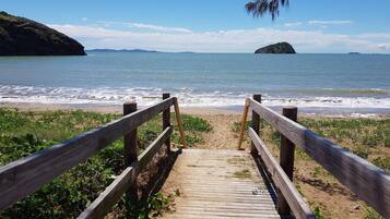 A wooden boardwalk leading to a beach with waves, a distant island, and a clear sky.