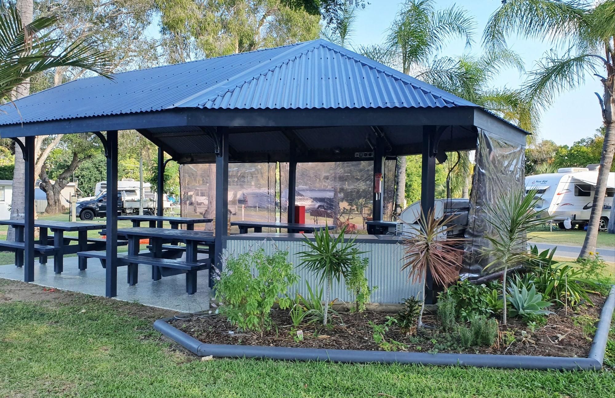 Un pabellón cubierto con bancos, rodeado de un jardín con diversas plantas y una palmera.