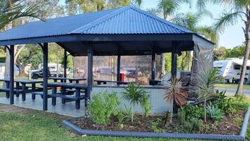 Un pabellón cubierto con bancos, rodeado de un jardín con diversas plantas y una palmera.