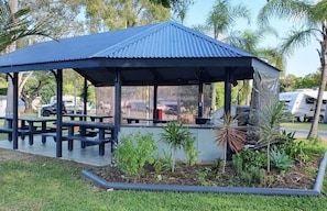 A covered pavilion with benches, surrounded by a garden with various plants and a palm tree.