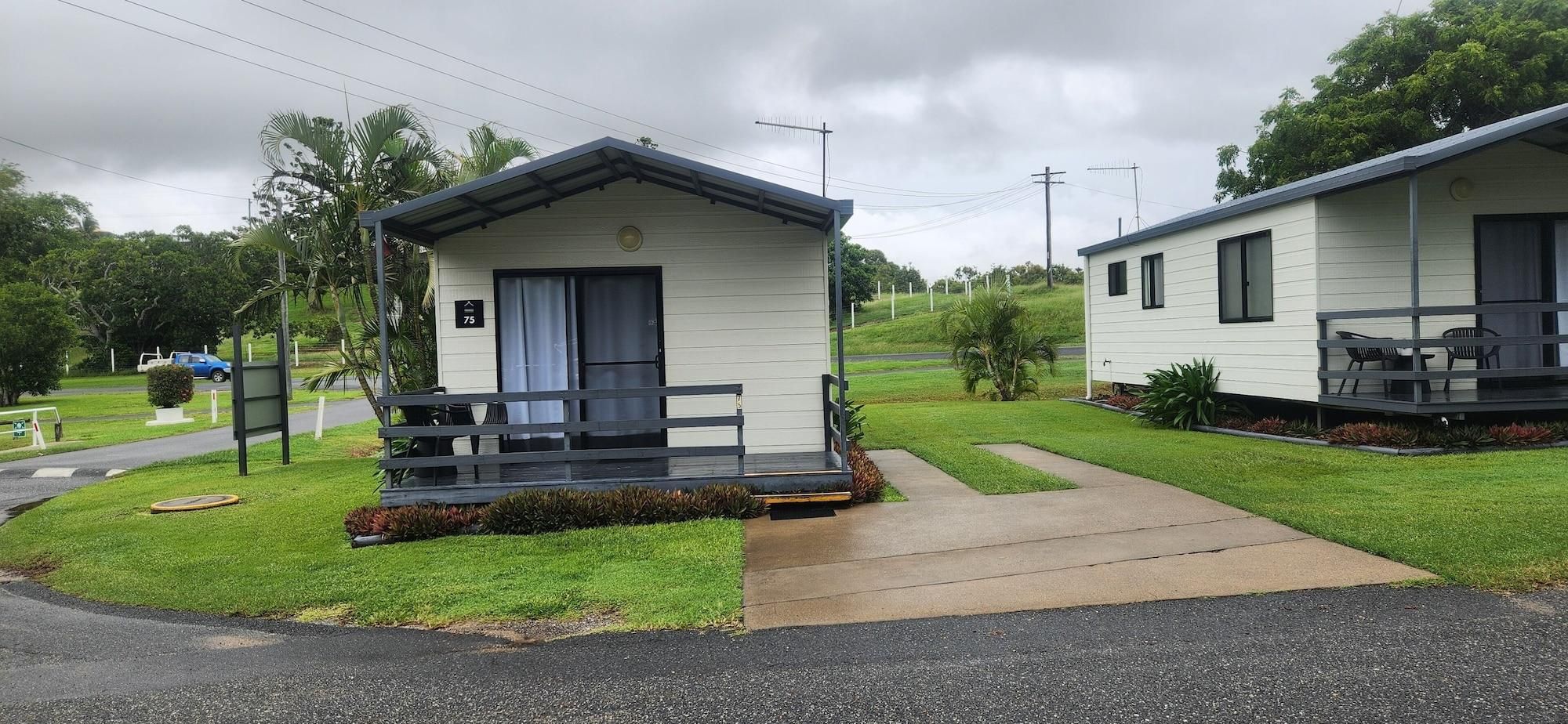 A row of small, single-story cabins with a porch and a small deck, surrounded by well-maintained grass and trees.
