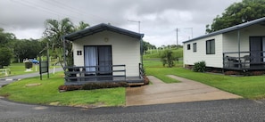 A row of small, single-story cabins with a porch and a small deck, surrounded by well-maintained grass and trees.