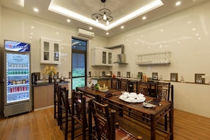 A modern kitchen with a dining area, featuring wooden furniture, a refrigerator stocked with beverages, and a large window providing natural light. - Mh Cherish Homestay (Hoa Lu)