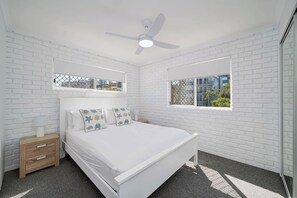 Een slaapkamer met een witte bakstenen muur, een bed met witte beddengoed, een nachtkastje, een lamp, een plafondventilator en een raam met uitzicht op gebouwen.