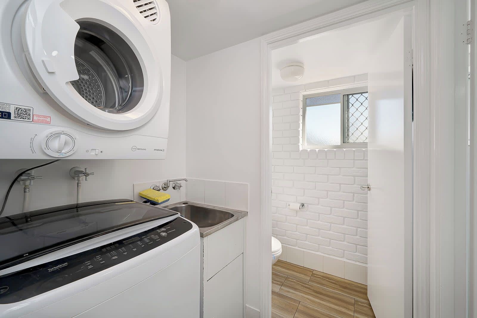 A compact laundry room with a front-loading washer and dryer, a small sink, and a window with a view of the outside.