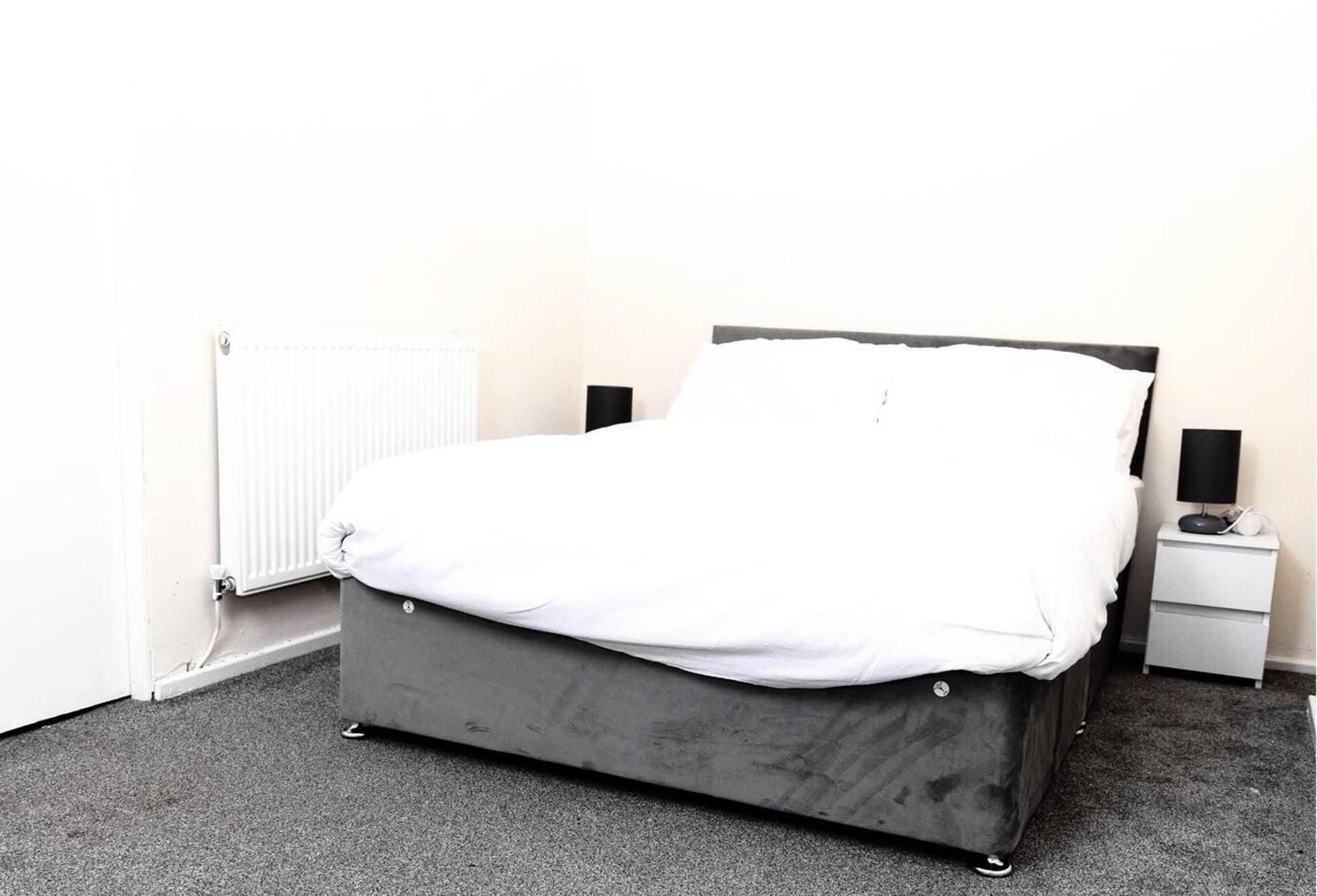 A single bed with a grey frame and white bedding in a room with a radiator and a nightstand with a lamp.