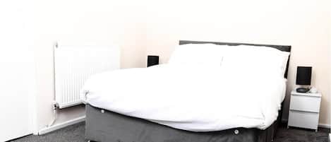 A single bed with a grey frame and white bedding in a room with a radiator and a nightstand with a lamp.