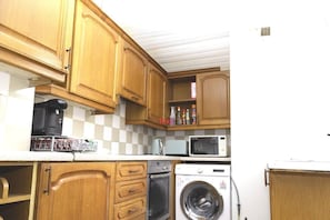 A compact kitchen with wooden cabinets, a coffee maker, a microwave, a washing machine, and a small shelf with various items.