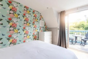 A bedroom with a floral wallpaper, a white bed, a dresser, and a balcony with outdoor furniture. - Creek View Cottage Little Petherick 2 Bedrooms (Wadebridge)