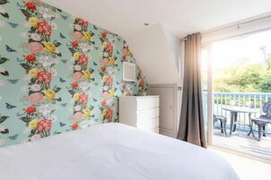 A bedroom with a floral wallpaper, a white bed, a dresser, and a balcony with outdoor furniture.