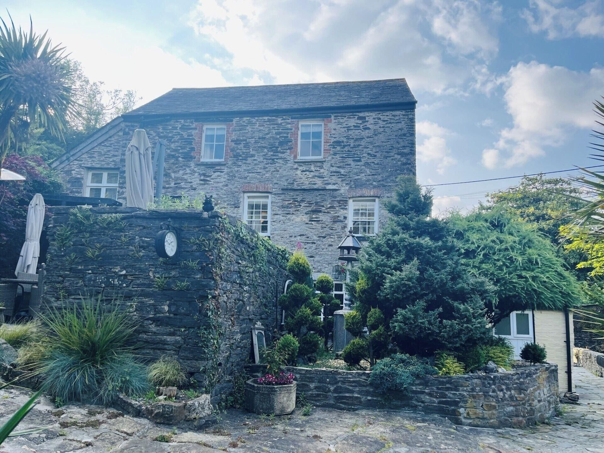 Ein Steinhaus mit geneigtem Dach, zahlreichen Fenstern und einem Garten mit verschiedenen Pflanzen und einem kleinen Brunnen.