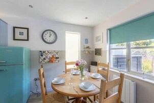 A dining area with a round table set for four, wooden chairs, a clock on the wall, and a window with a view of the outside.