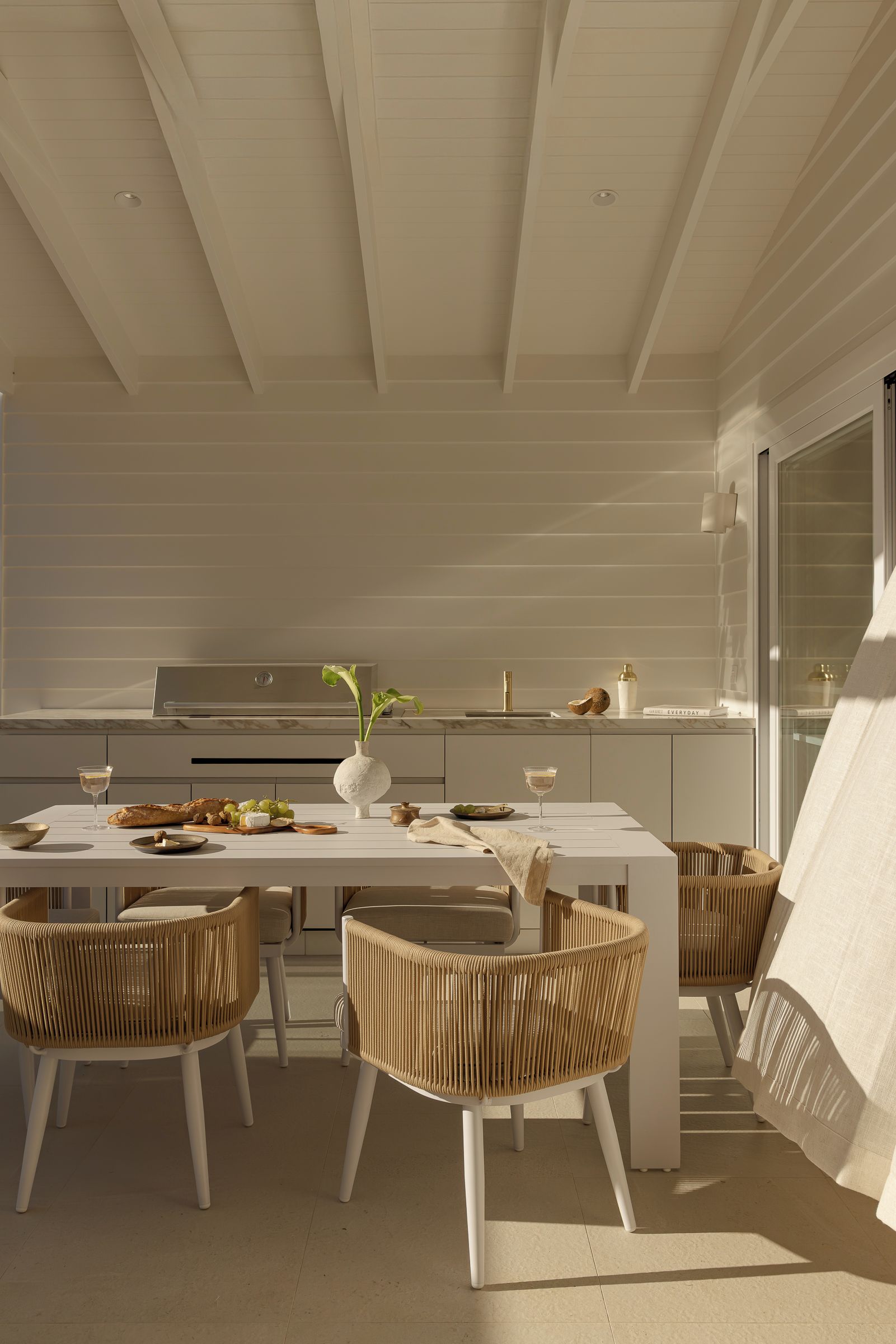 A modern kitchen with a dining area, featuring a white table, wicker chairs, and a vase with greenery.