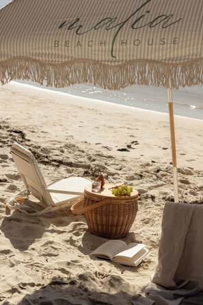 Ein Liegestuhl, ein Korbwägelchen mit Trauben, ein Buch und ein Strandsonnenschirm am Sandstrand.