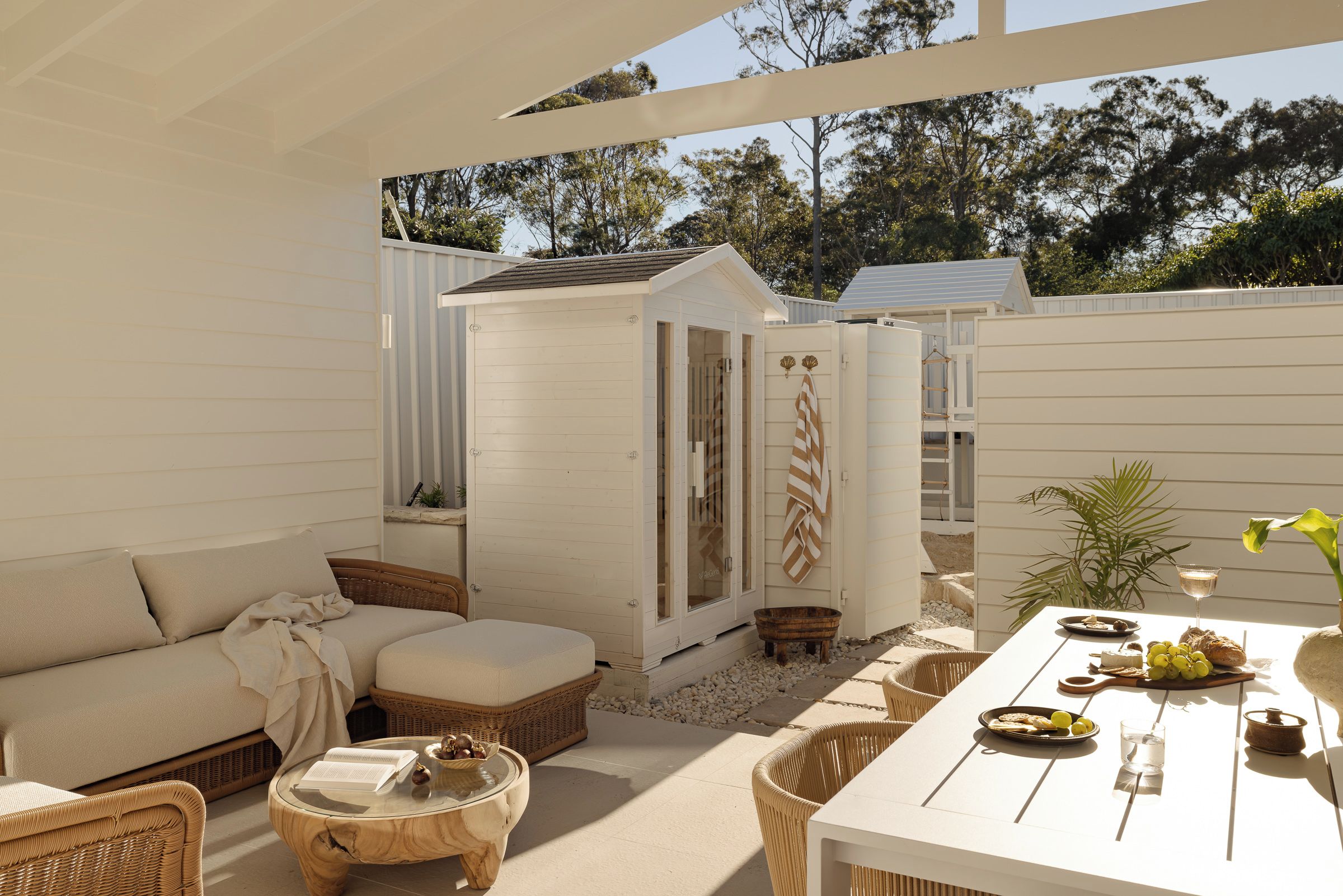 A covered outdoor patio with a white sofa, wicker furniture, a small wooden structure, and a dining table set with food and drinks.