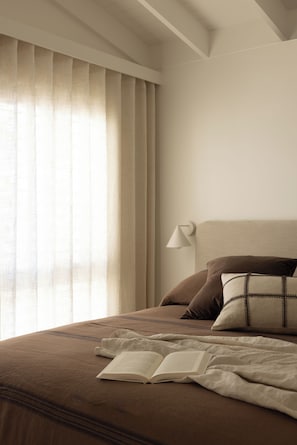 Cama bem arrumada com um livro, um travesseiro e uma luminária de cabeceira em um quarto com janela e cortinas.
