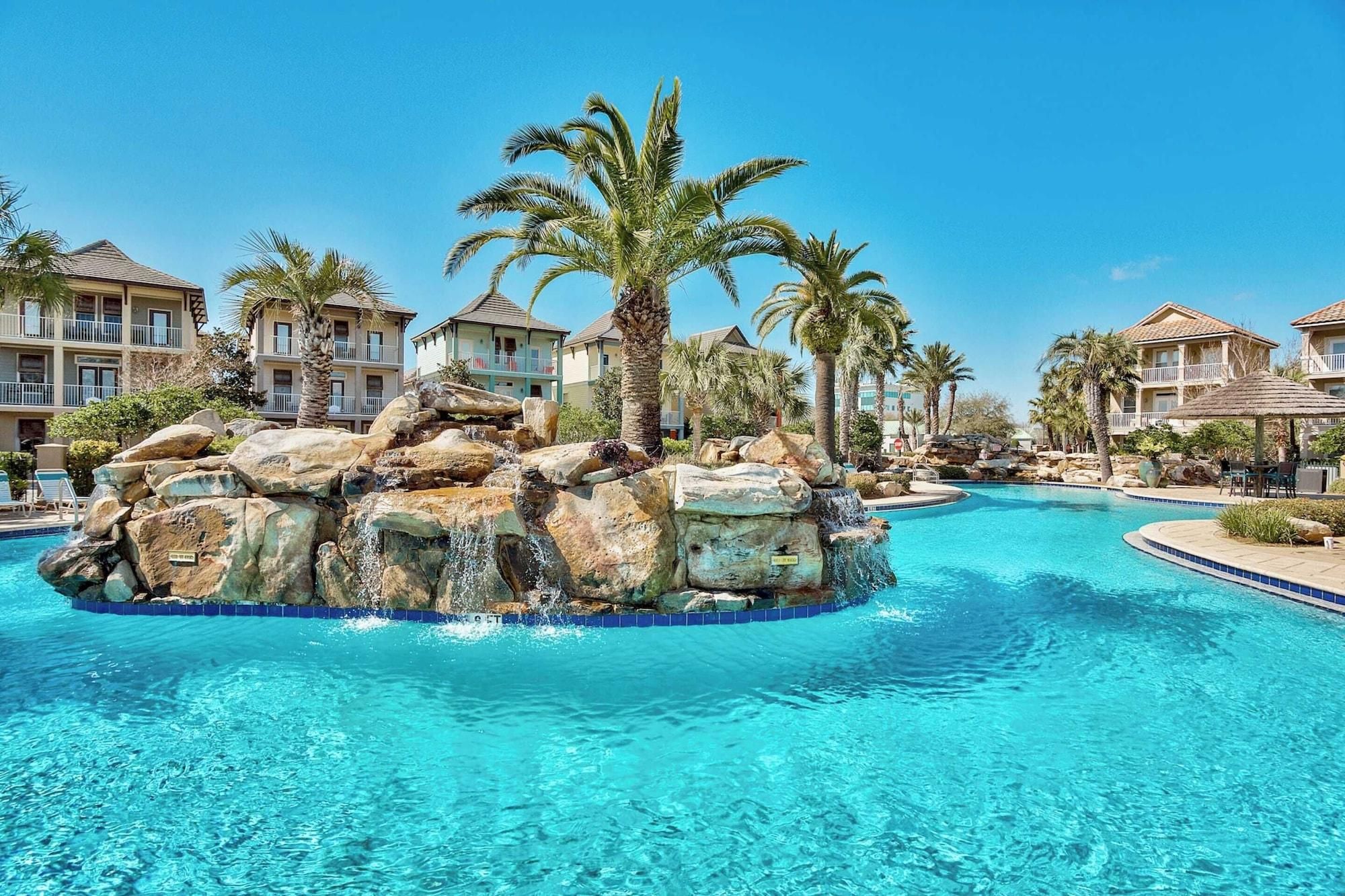 A resort pool with a rocky island and palm trees, surrounded by hotel buildings.