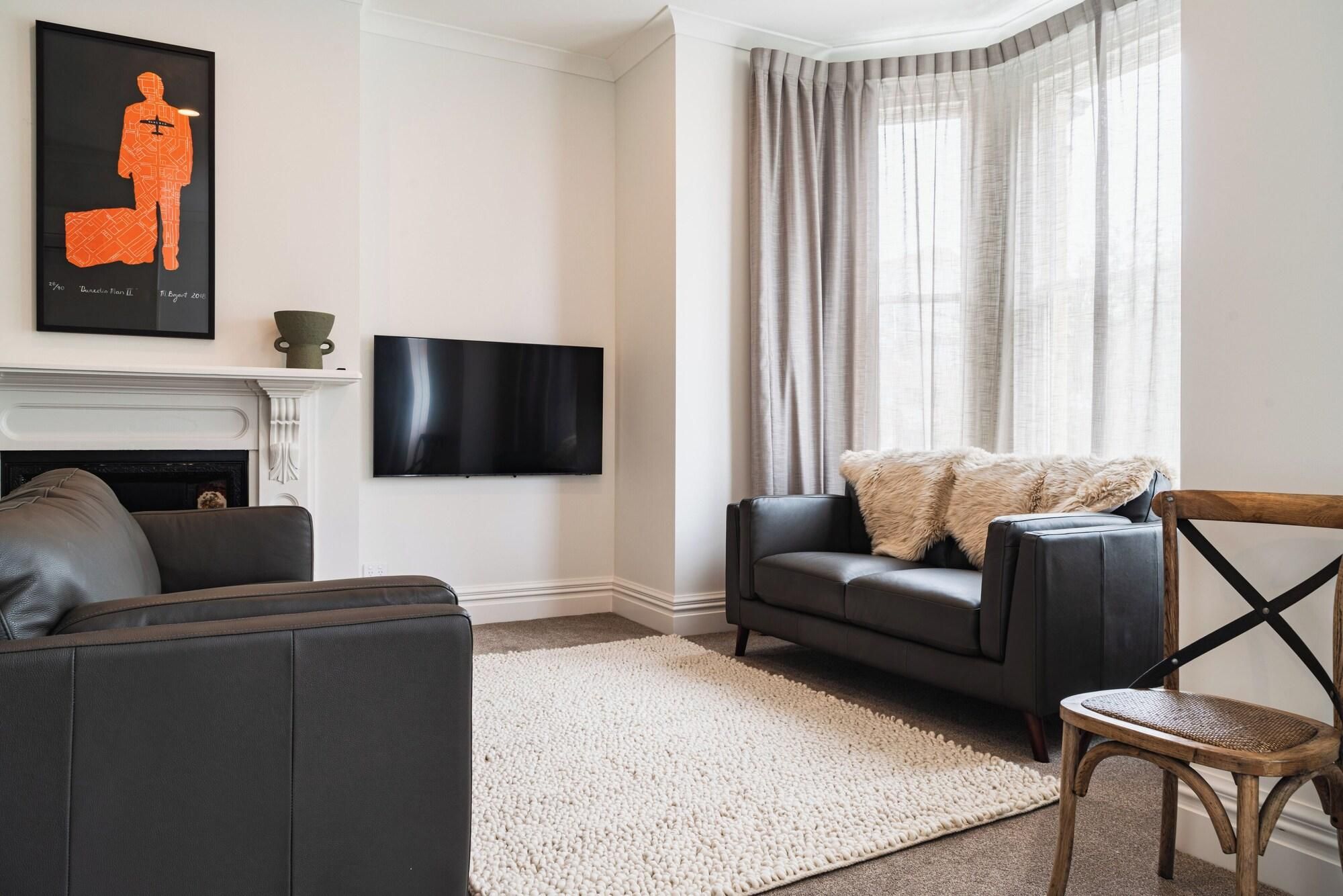 A living room with a fireplace, a flat-screen TV mounted on the wall, a grey sofa, a white rug, and a wooden chair.