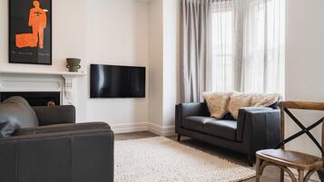 A living room with a fireplace, a flat-screen TV mounted on the wall, a grey sofa, a white rug, and a wooden chair.
