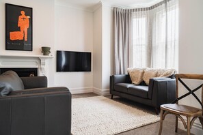 Ein Wohnzimmer mit Kamin, einem an der Wand befestigten Flachbildfernseher, einem grauen Sofa, einem weißen Teppich und einem Holzsessel.