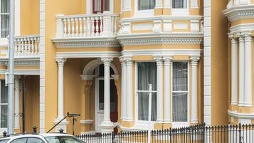 A two-story building with a balcony, a car parked in front, and a black iron fence.