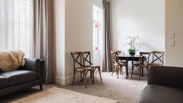 Una sala de estar con un sofá, una mesa de centro y una mesa de comedor con sillas. Hay una ventana con cortinas, una alfombra y una planta en maceta.