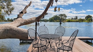A wooden deck with a round table and four chairs, overlooking a lake with a fallen tree branch and hanging lanterns.