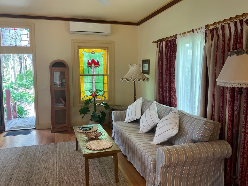A living room with a sofa, a table with a woven placemat, a potted plant, a stained glass window, and a door leading outside.