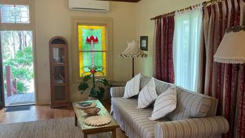A living room with a sofa, a table with a woven placemat, a potted plant, a stained glass window, and a door leading outside.