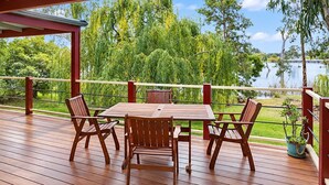 A wooden deck with a table and chairs, overlooking a lake with trees and a clear sky.