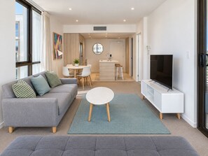 A modern living room with a grey sofa, a TV, and a dining area with a round table and chairs. - City Dreams at Avani | Chic High-Rise Stay (Broadbeach)