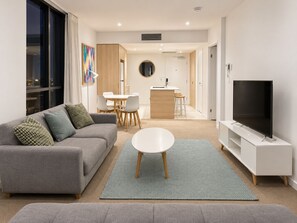 A modern living room with a grey sofa, a round coffee table, and a flat-screen TV on a white stand. - City Dreams at Avani | Chic High-Rise Stay (Broadbeach)