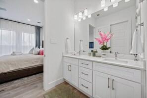 A modern hotel room with a large bed, a bathroom with a vanity and mirror, and a view of the outdoors. - Modern 2BR/2.5BA Townhome | 2-Car Garage (Dallas)
