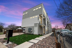 A modern two-story building with a flat roof, a visible garage door, and a well-maintained lawn with stepping stones leading to the entrance. - Modern 2BR/2.5BA Townhome | 2-Car Garage (Dallas)