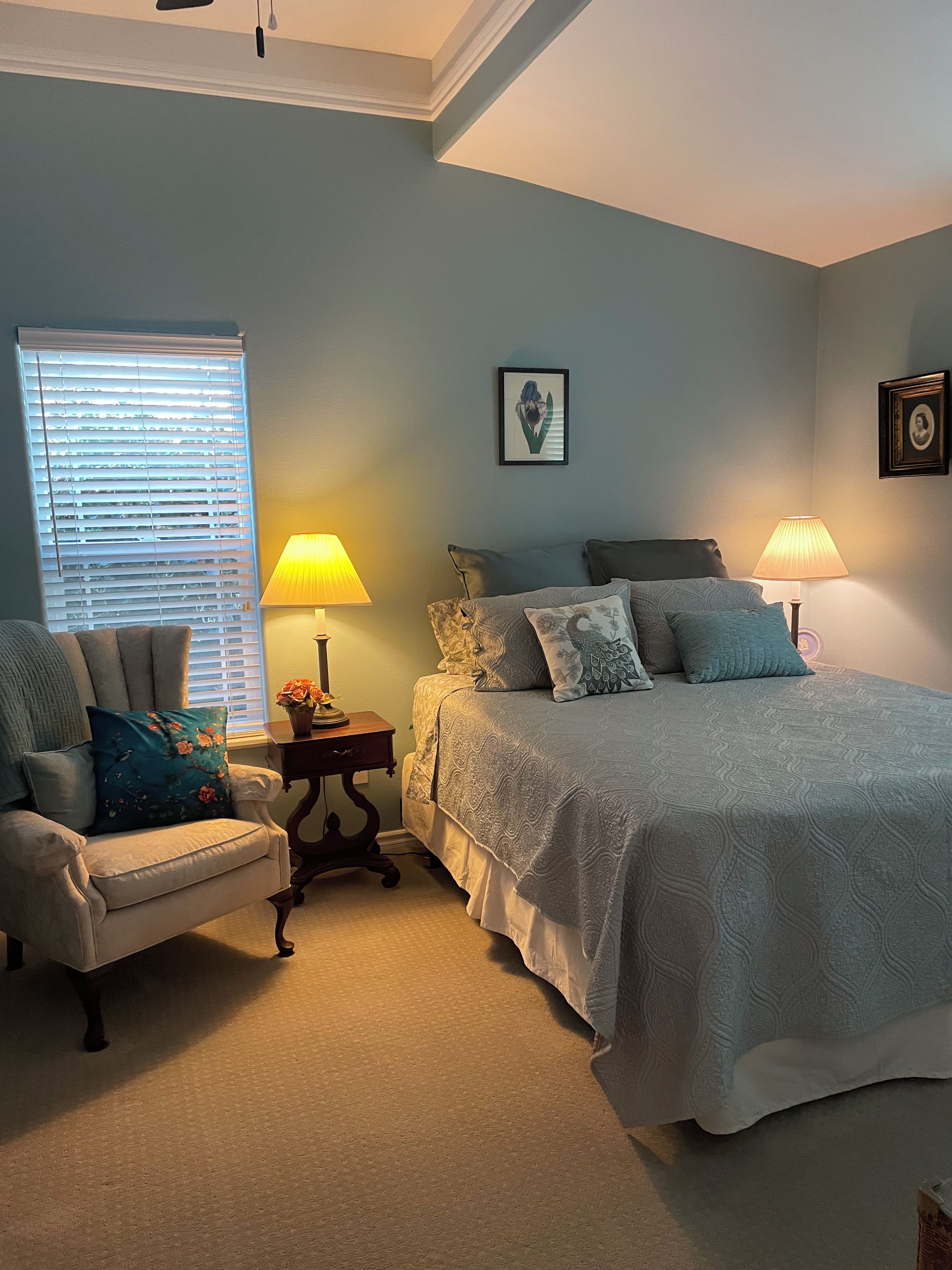 A bedroom with a bed, a chair, two lamps, a picture on the wall, and a window with blinds.