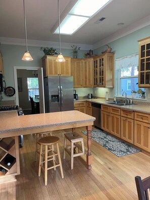 A kitchen with wooden cabinets, a stainless steel refrigerator, a large island with stools, and a window with lace curtains.