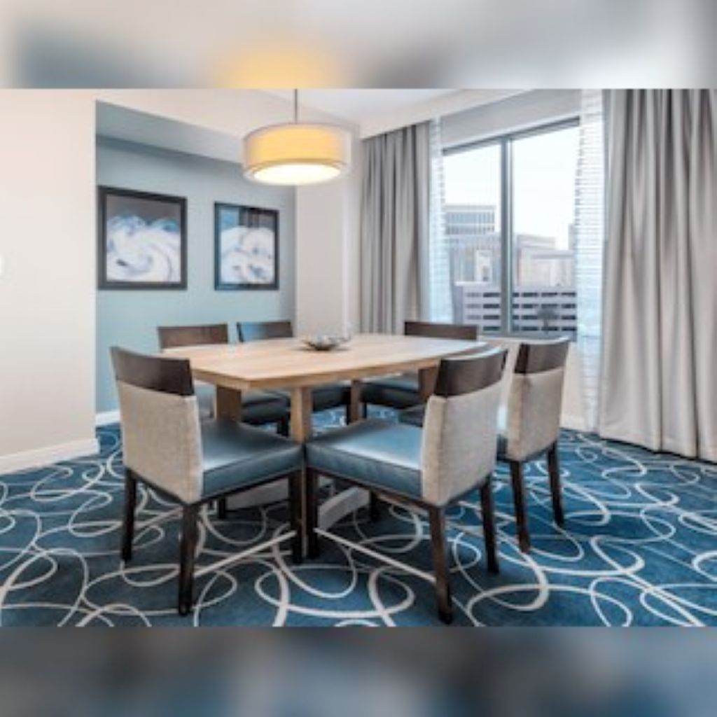 Une chambre d’hôtel équipée d’une table à manger, de chaises, d’une grande fenêtre avec des rideaux et d’un tapis à motifs.
