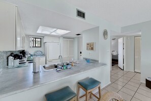 A modern kitchen with a central island, a sink, and bar stools. There is a window above the sink and a clock on the wall. - Coastal Condo | Pool View + Heated Pool (St. Augustine)