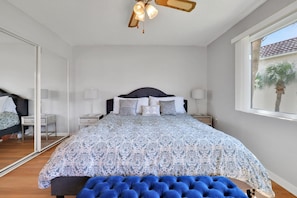 A bedroom with a large bed, bedside tables, a ceiling fan, and a window with a view of a palm tree. - Coastal Condo | Pool View + Heated Pool (St. Augustine)