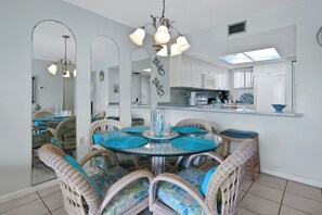 A dining area with wicker chairs and a glass table, a kitchen with white cabinets, and a skylight. - Coastal Condo | Pool View + Heated Pool (St. Augustine)
