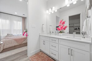 Ein modernes Schlafzimmer mit einem großen Bett, ein Badezimmer mit weißem Waschtisch und Spiegel sowie ein Blick nach draußen.