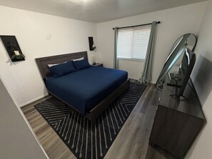 Ein Schlafzimmer mit einem Bett, einem Nachttisch, einem Spiegel, einem Fenster mit Jalousien und einem an der Wand befestigten Regal.