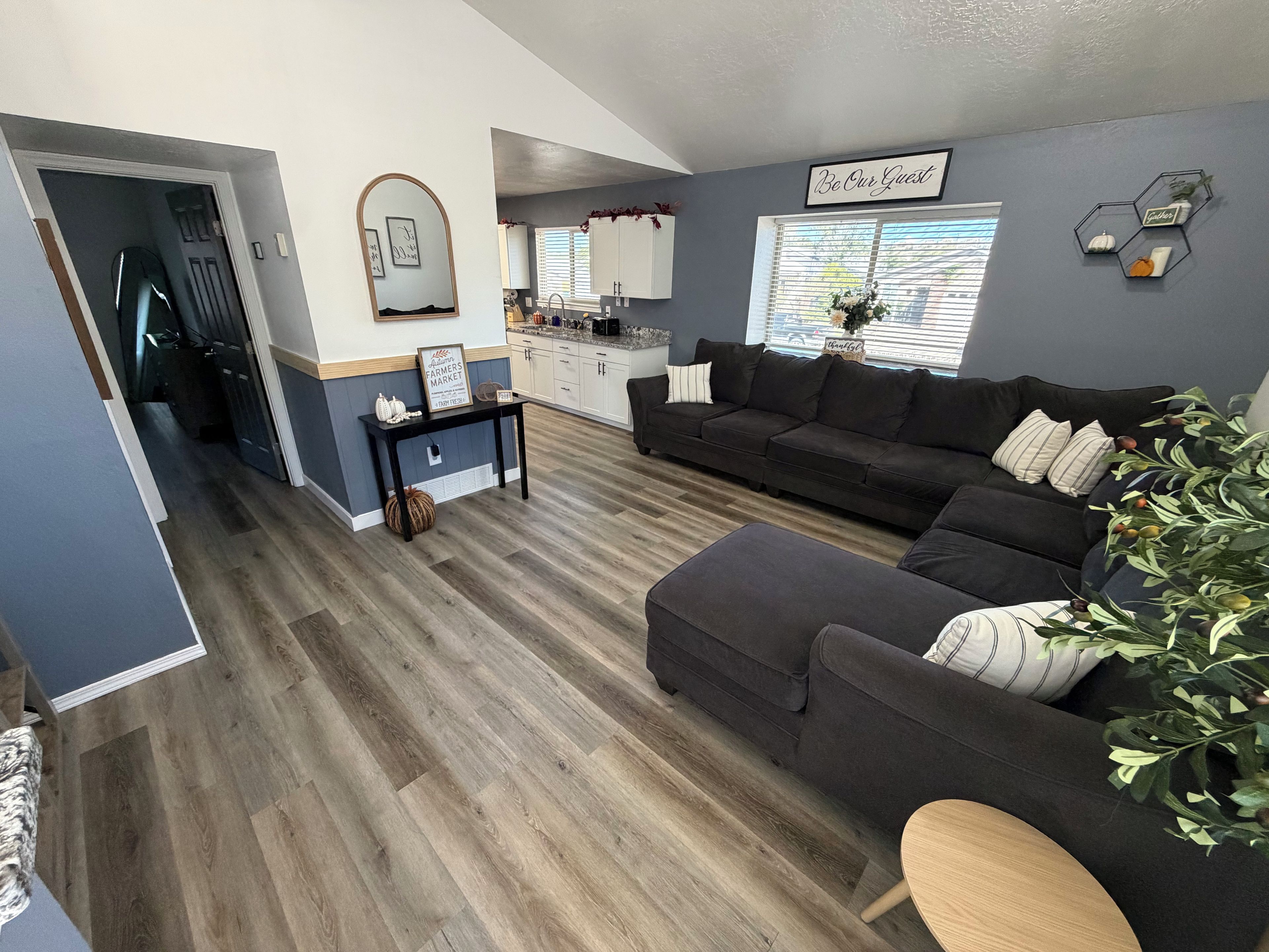 A modern living room with a dark grey sofa, a wooden coffee table, and a kitchen in the background.